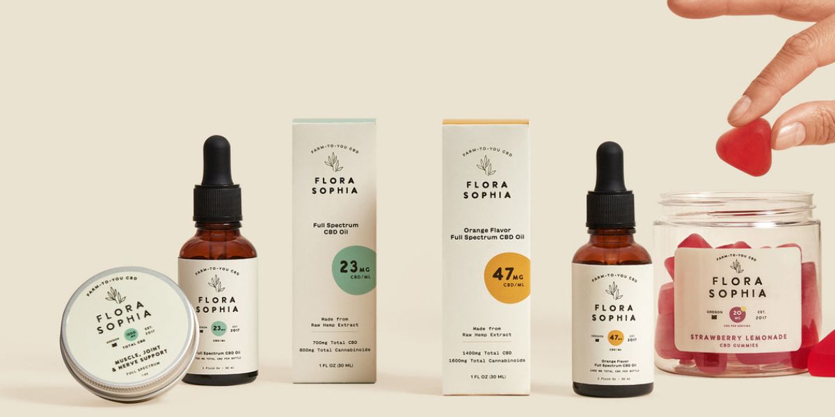 Flora Sophia tinctures, salve and gummies lined up on a cream table, with a hand picking a red gummy from the jar.