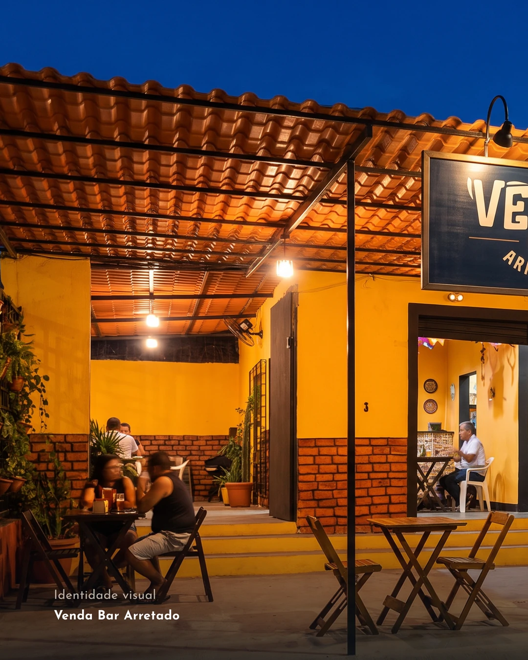 Fachada amarela do Venda Bar Arretado à noite, com a placa sob o pergolado e mesas de madeira